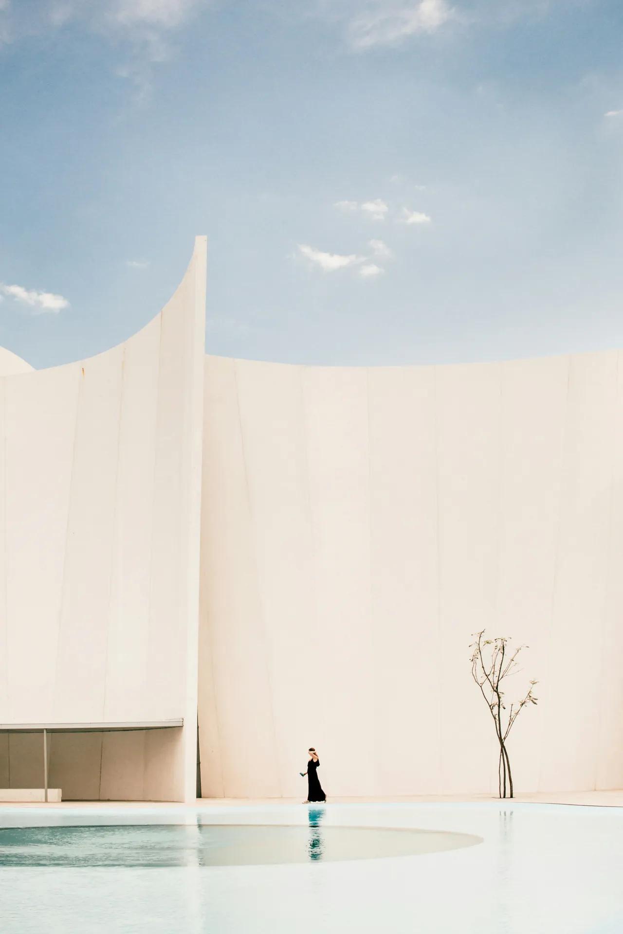 A figure in black before a curved white wall, mirrored in a shallow pool