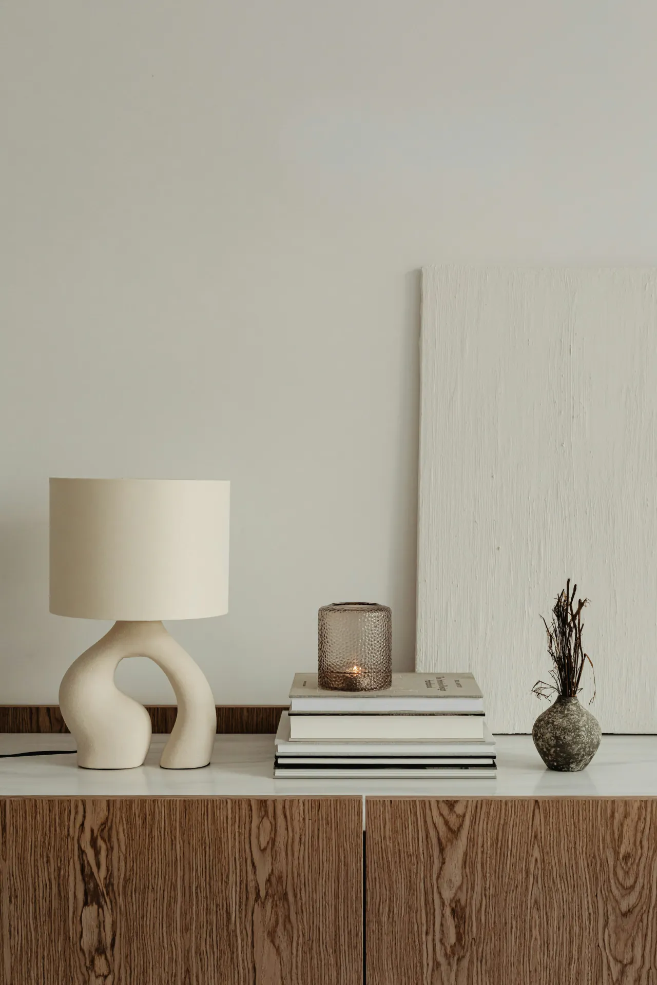 A ceramic lamp, a stack of books and a small vase on a wooden sideboard